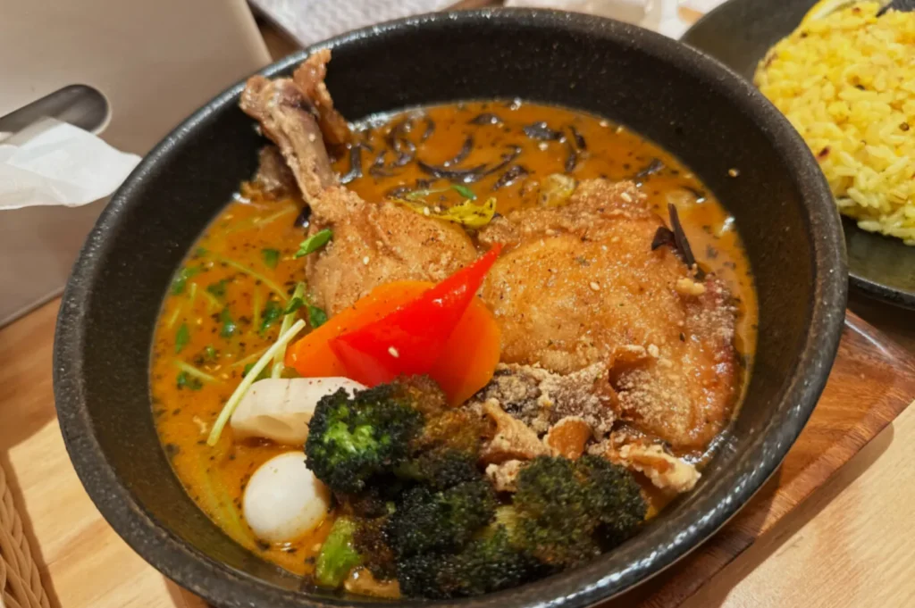 A black bowl filled with curry soup, featuring chicken, broccoli, mushrooms, red bell pepper, and herbs. A plate of yellow rice is nearby. Cozy and appetizing.