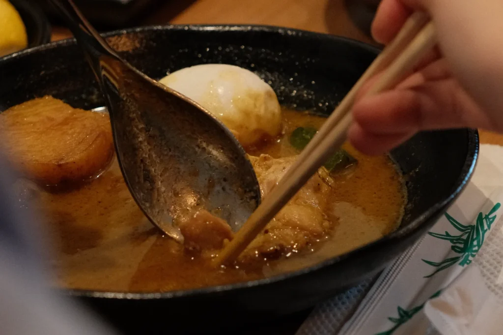 A hand using chopsticks to lift a piece of meat from a black bowl of soup, which appears to contain an egg and other ingredients.