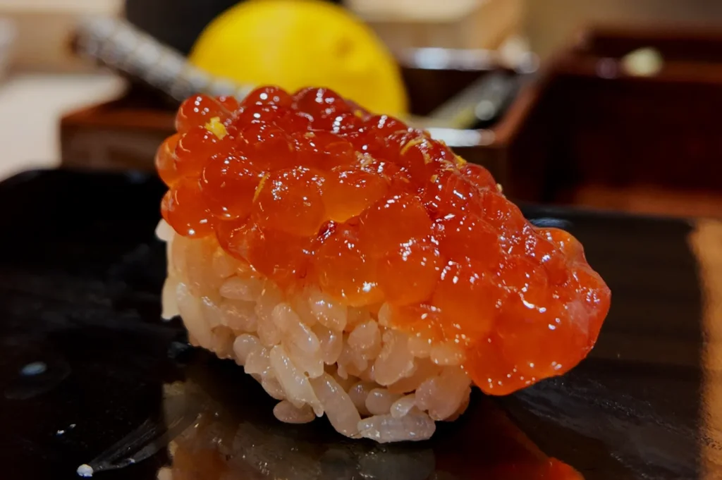 Close-up of ikura sushi with shiny, orange salmon roe atop glossy rice on a dark plate. In the blurred background, a yellow lemon is visible. Fresh and appetizing.