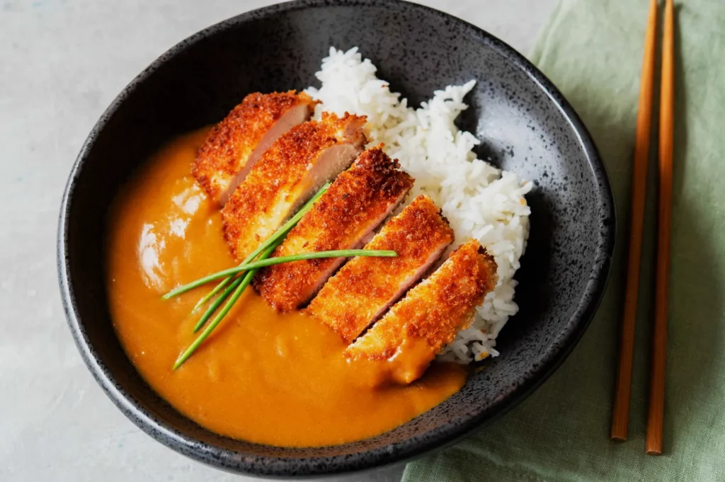 A black bowl with crispy breaded chicken slices on white rice, topped with golden curry sauce. Garnished with green chives. Chopsticks on the side.