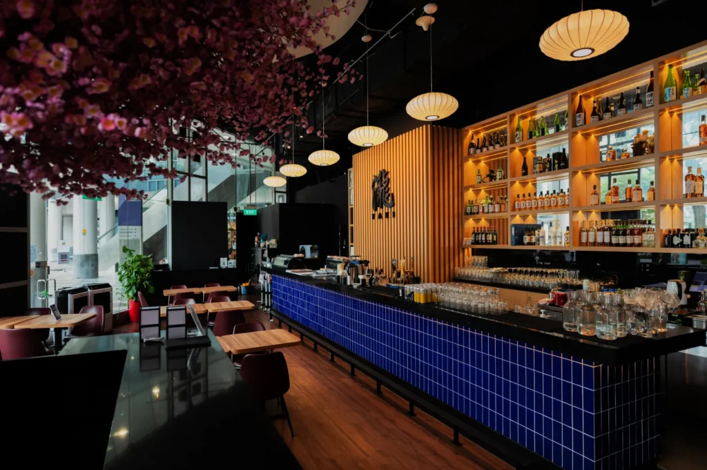 Modern cafe interior with a long bar featuring blue tiles, wooden shelves stocked with bottles, and hanging lanterns. Cherry blossom decor adds elegance.