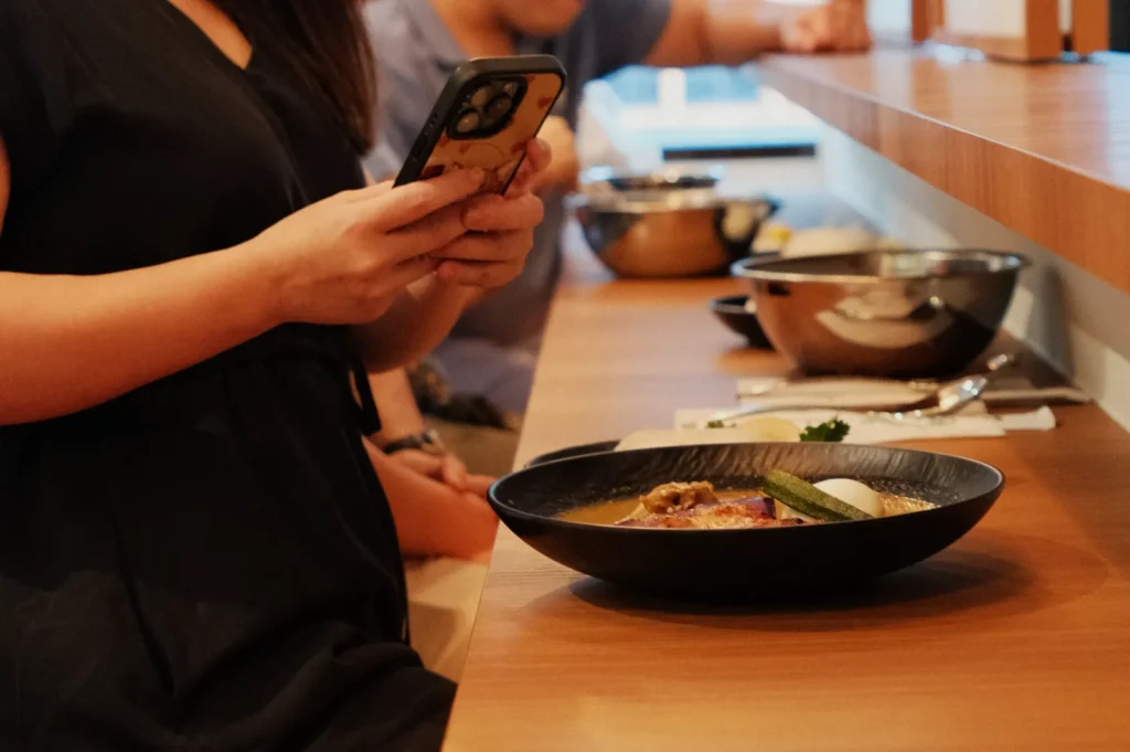 A person sitting at a table with a bowl of noodles, holding a phone, while others are seated in the background.