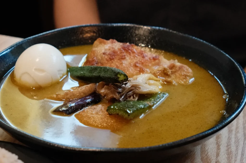 A bowl of rich yellow curry with pieces of chicken, vegetables, and a poached egg placed in a black bowl on top of a wooden dining table with a person behind.