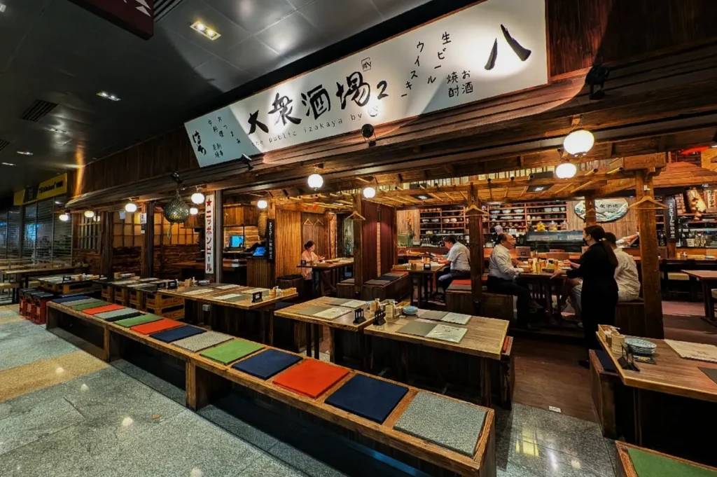 Cozy Japanese izakaya with wooden decor, colorful seat cushions, and patrons seated inside. Warm lighting creates an inviting atmosphere.