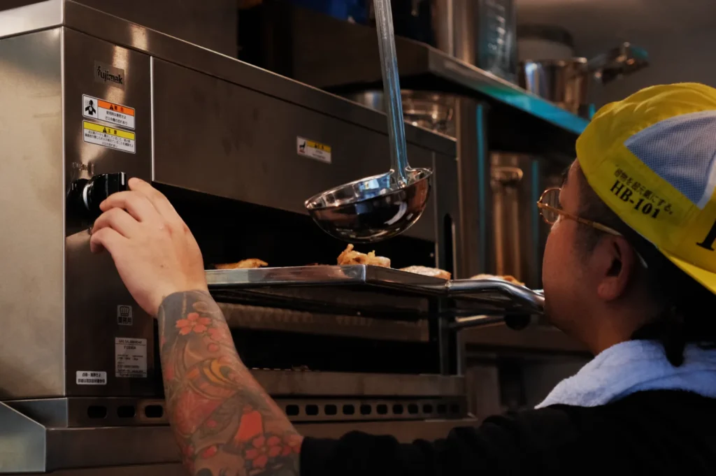 A person with tattoos is using a ladle to take food out of an oven in a kitchen setting. They are wearing a yellow cap and a towel around their neck.