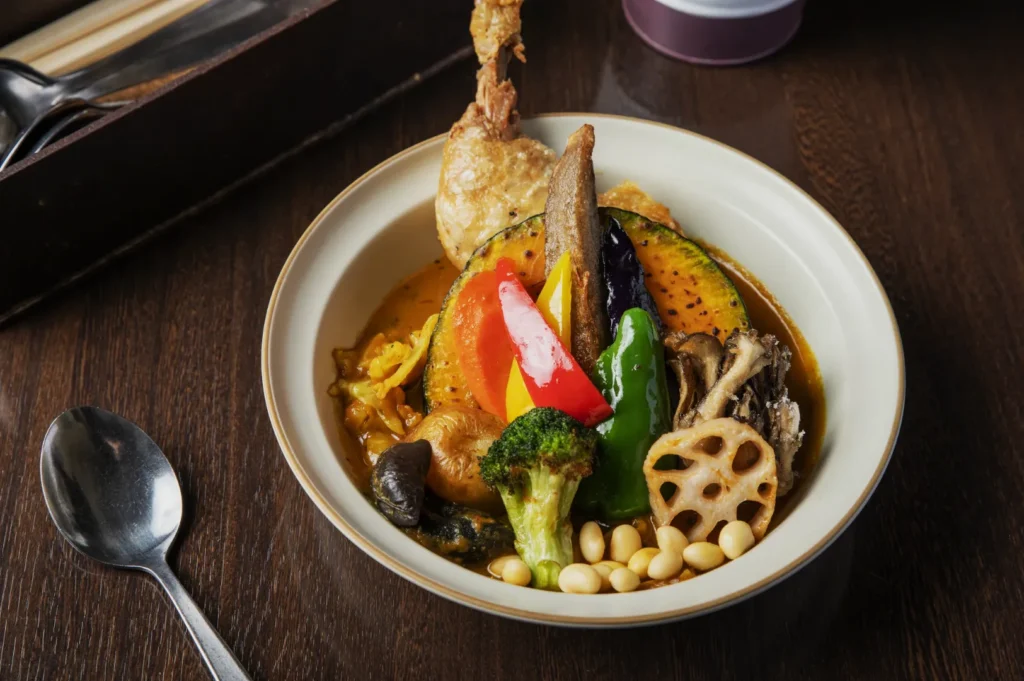 A bowl of vibrant vegetable curry against a wooden table backdrop. Colorful ingredients include squash, bell peppers, lotus root, broccoli, and soybeans, with fried chicken.
