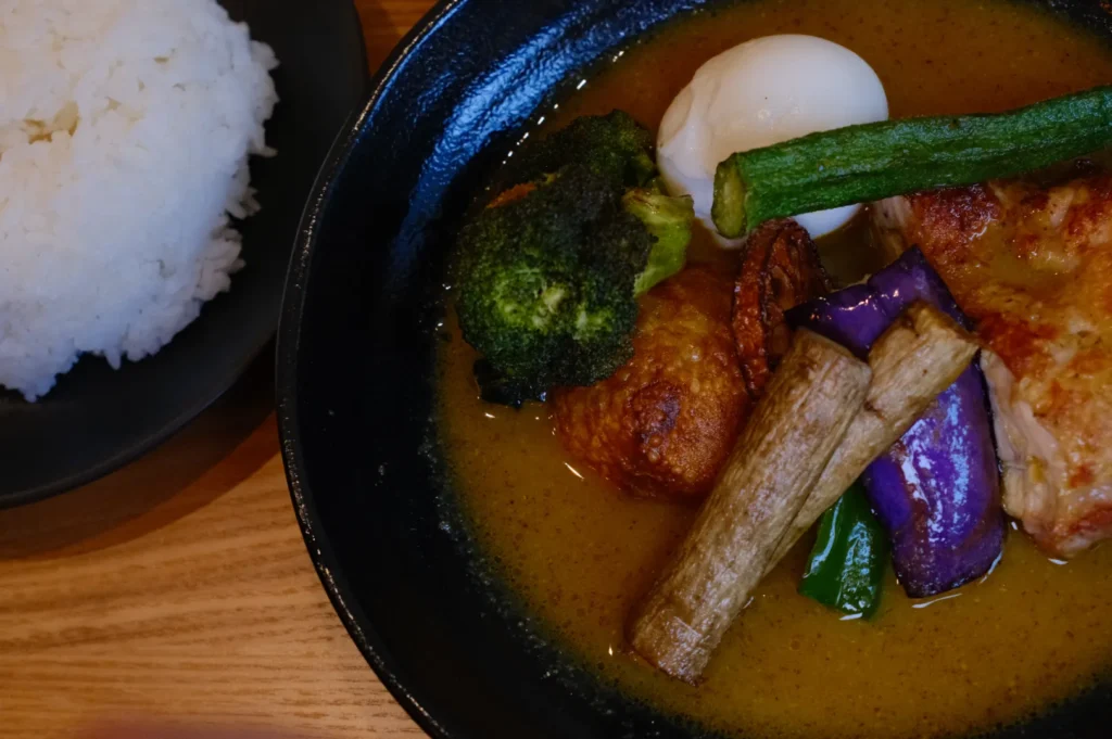 A bowl of soup curry with various vegetables and a piece of chicken, served with a side of white rice. Ecah placed in black bowls on a wooden table.