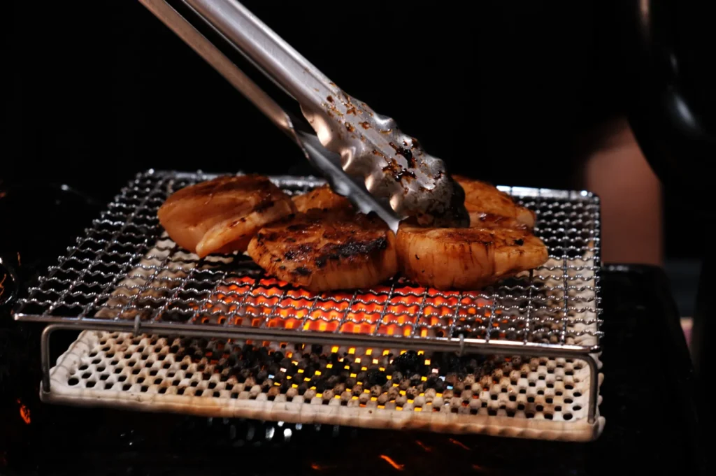 Grilled scallops on a metal mesh over glowing charcoal with tongs flipping one piece. The scene conveys a warm, appetizing atmosphere.