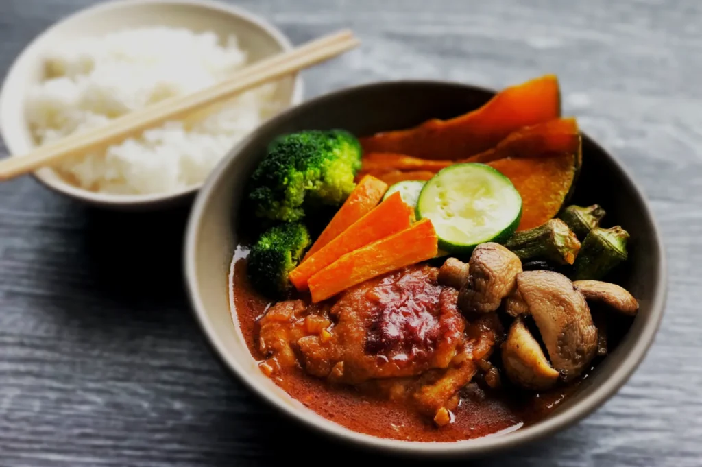 A bowl of vibrant curry with mushrooms, carrots, zucchini, broccoli, and pumpkin slices in a rich sauce, paired with a bowl of white rice and chopsticks.