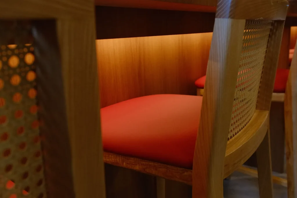 Wooden chairs with red cushions are lined up under a counter, illuminated by soft warm lighting, creating a cozy and inviting ambiance.