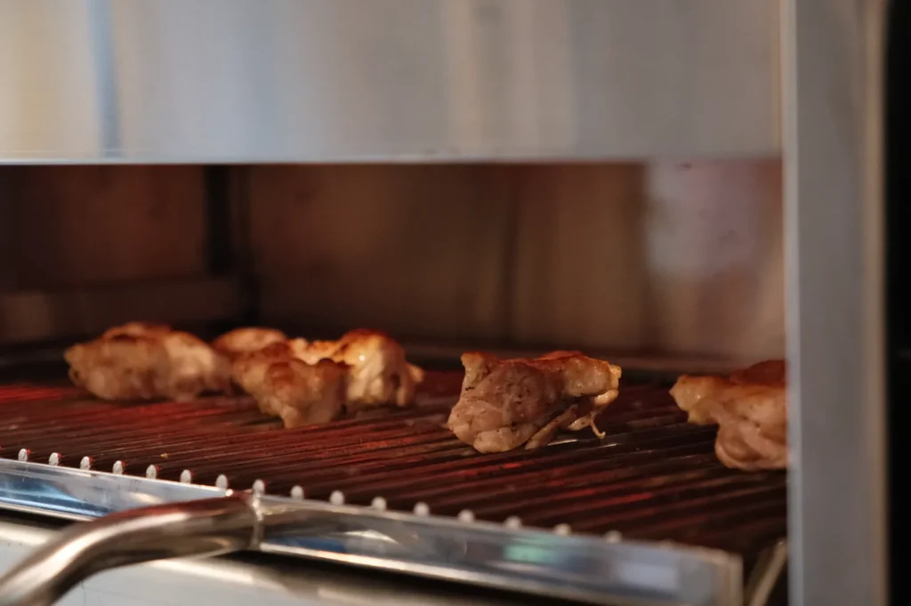 Four chicken thighs evenly placed apart, grilling on an indoor barbecue. Low heat rises from the barbecue, metal parts of the kitchen framing everything.