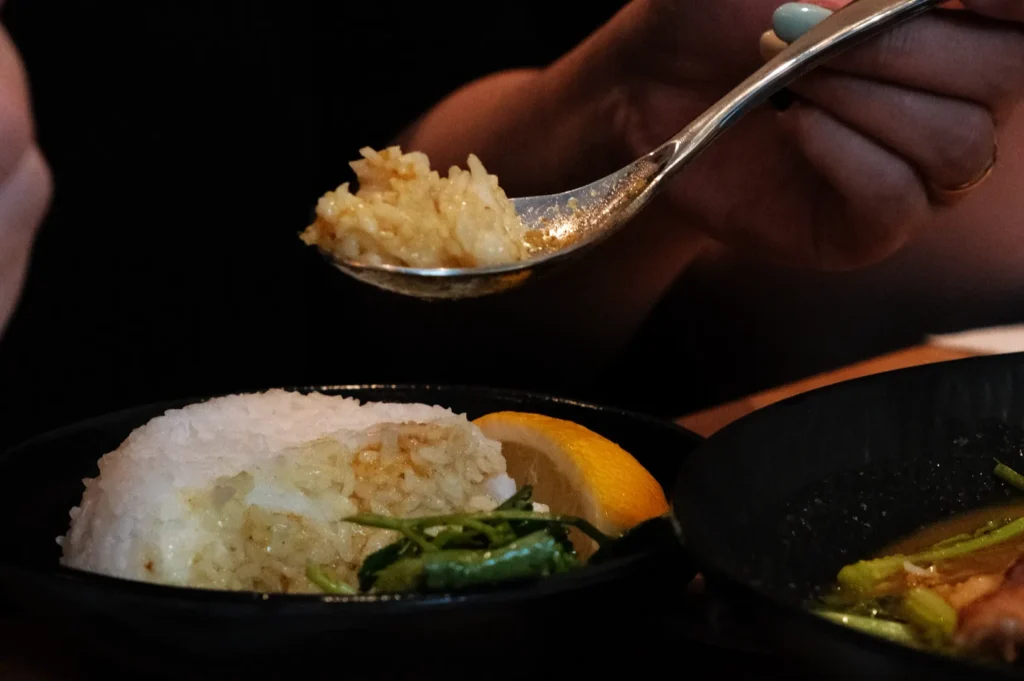 A hand holding a spoon with rice, near a plate of rice and greens garnished with lemon.