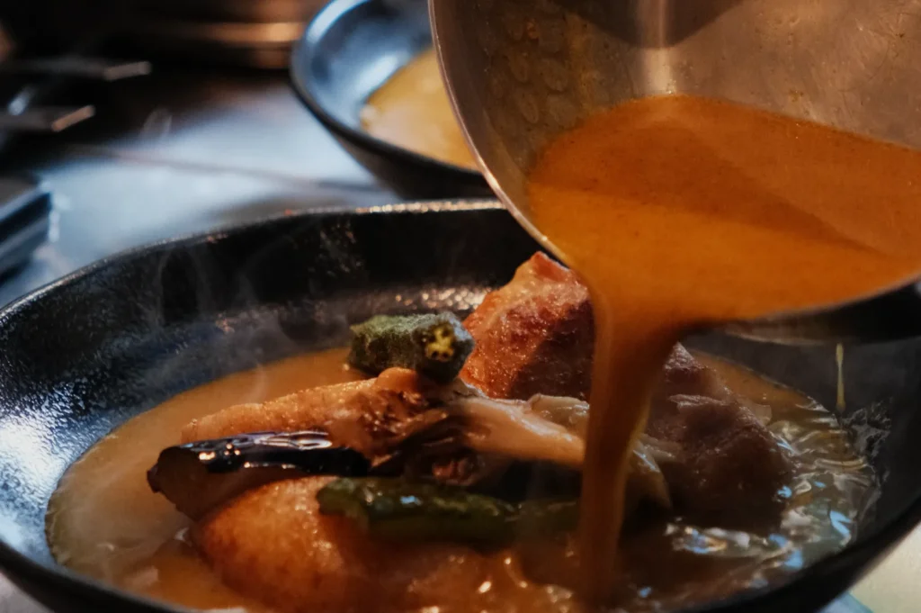 A pot of sauce being poured over a dish containing vegetables and possibly meat in a black bowl.