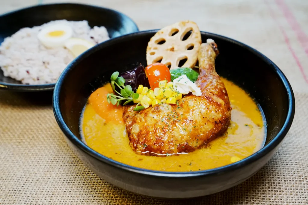 A black bowl of vibrant curry with grilled chicken, assorted vegetables, corn, and a lotus root slice. In the background, a bowl of rice with sliced eggs.