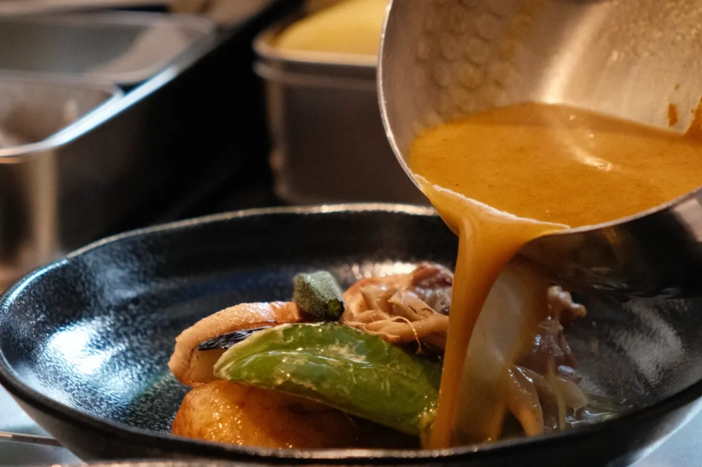 A close-up of Chef Masa pouring sauce over a dish in a black bowl, featuring various ingredients like chicken and vegetables.