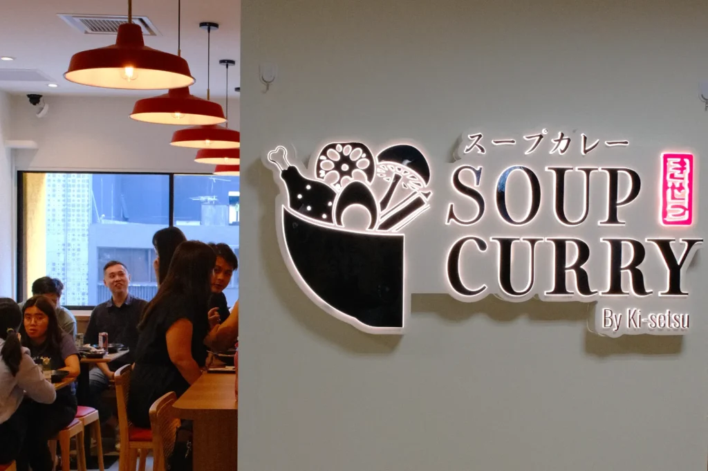 A restaurant interior featuring a sign that reads "Soup Curry" in both Japanese and English, with a bowl graphic and patrons dining in the background.