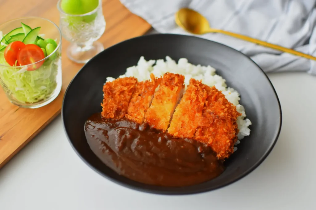 A dish of golden-brown katsu curry served over white rice on a black plate, with a side of fresh salad featuring lettuce, cucumber, and cherry tomatoes in a glass.