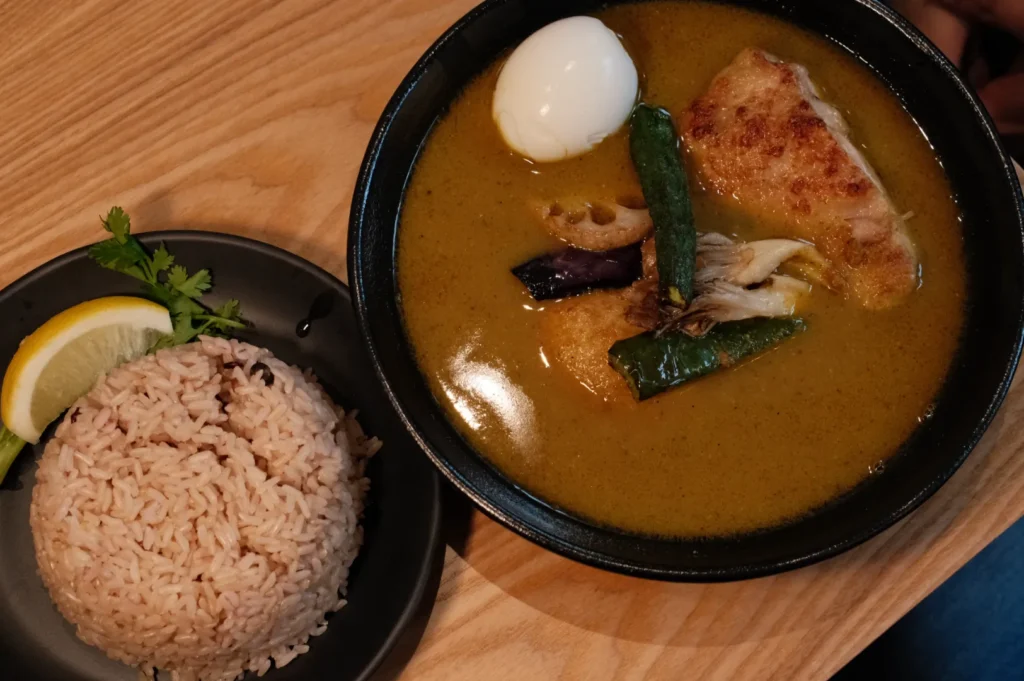 A bowl of curry soup with chicken, a boiled egg, and vegetables sits beside a plate of rice garnished with lemon and parsley, on a wooden table.