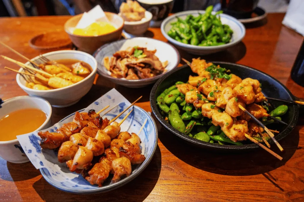 A table of Japanese skewers, edamame, simmered dishes, and tea in a cozy setting, conveying warmth and a sense of shared enjoyment.