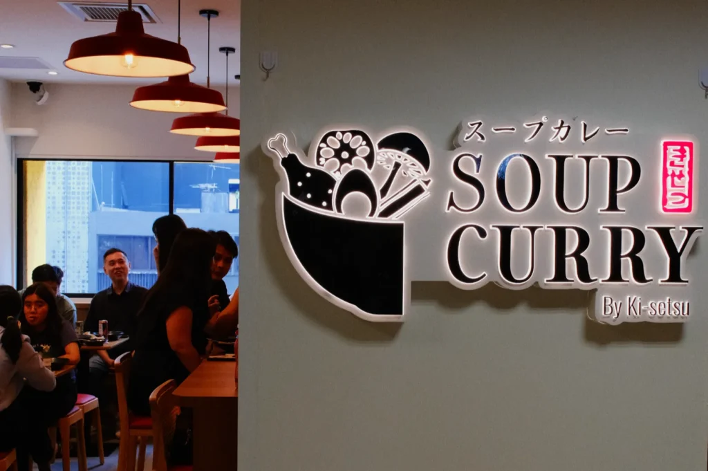 A restaurant interior featuring a neon sign that reads "SOUP CURRY By K-sotsu" in English and Japanese, with patrons seated at tables.