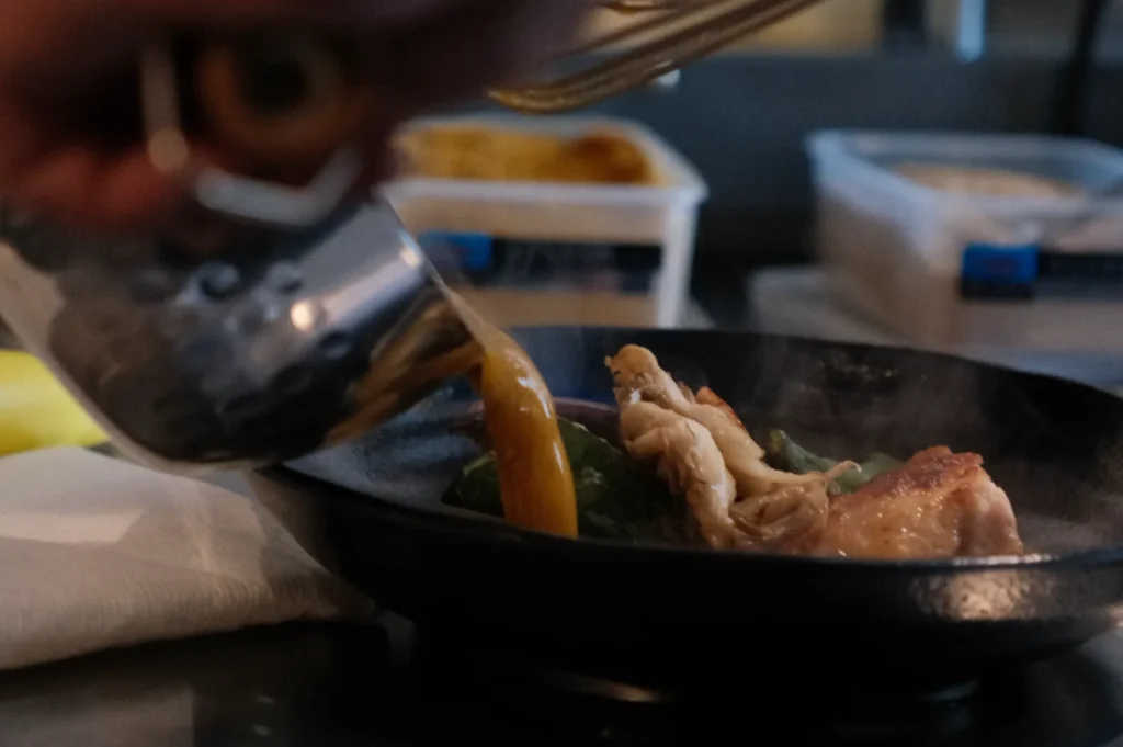 A person pouring sauce over a sizzling dish in a skillet, with steam rising and ingredients like chicken and vegetables visible.