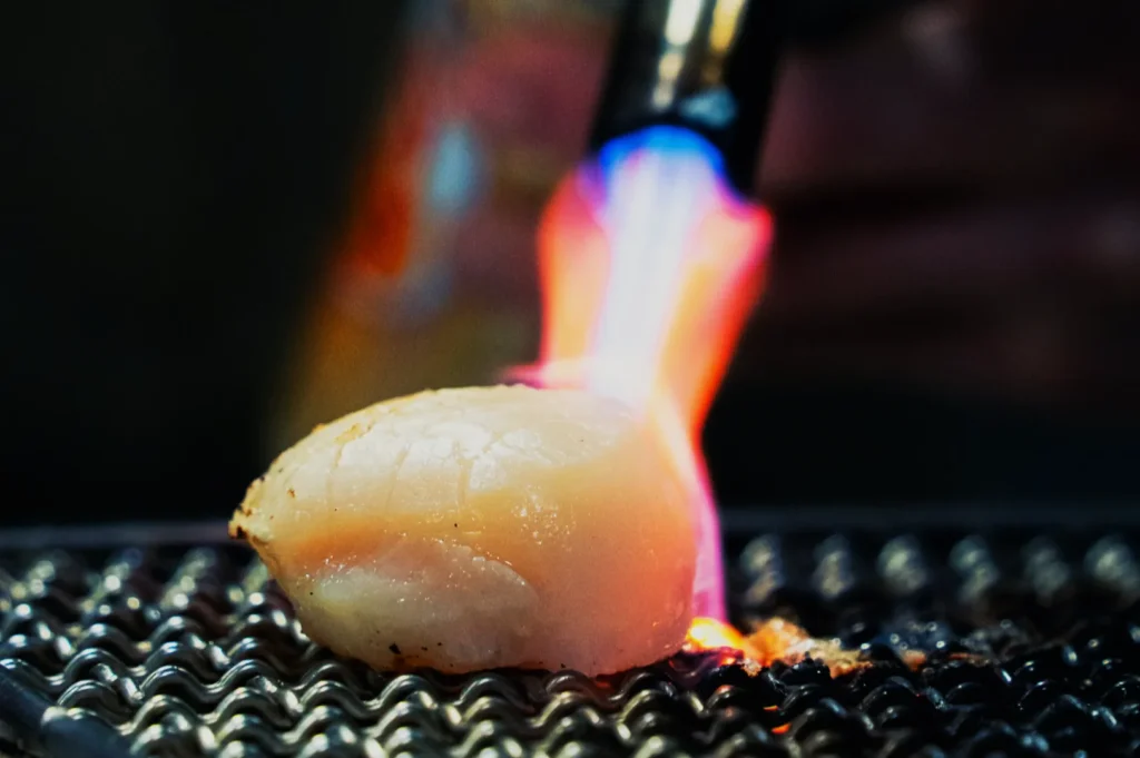 A scallop being torched on a grill, with blue and orange flames creating a caramelized surface. The setting is warm and culinary-focused.