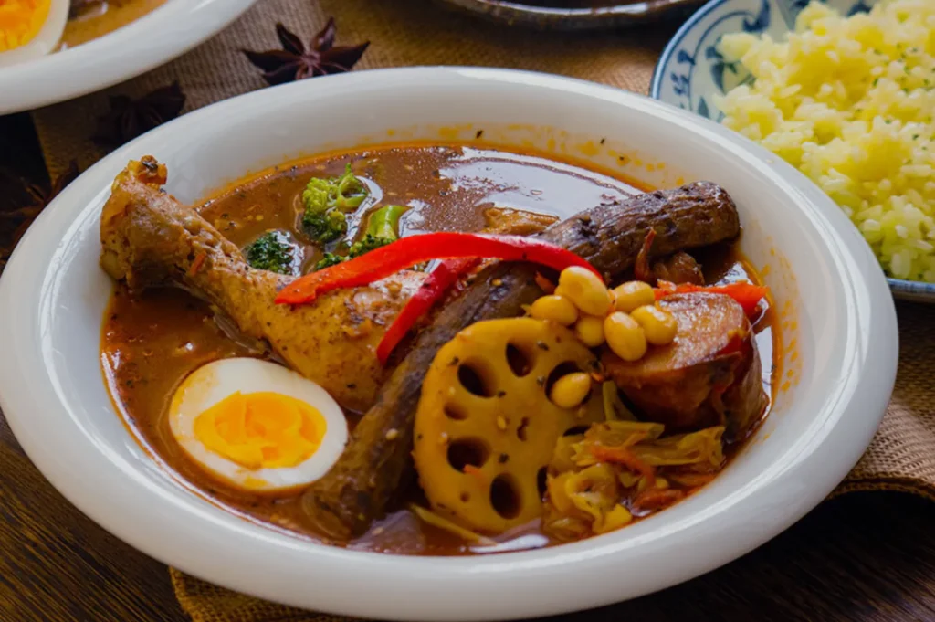 A bowl of Japanese curry soup with a chicken drumstick, boiled egg, lotus root, red pepper slices, and vegetables, next to a plate of yellow rice.