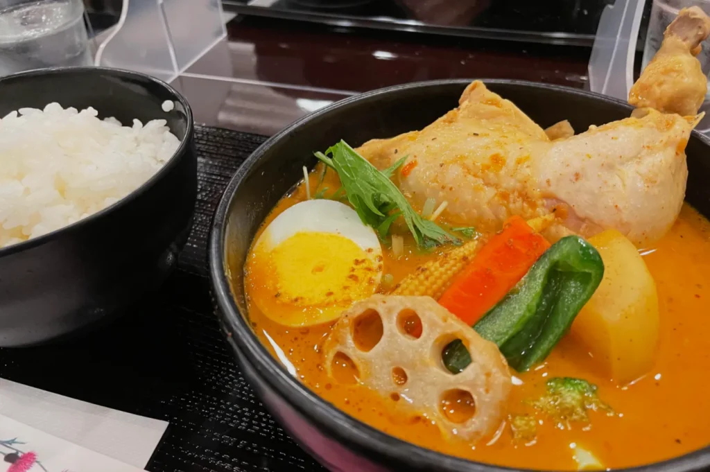 A bowl of vibrant orange curry with a chicken leg, colorful vegetables, and a boiled egg, served alongside a bowl of white rice. Inviting and comforting.