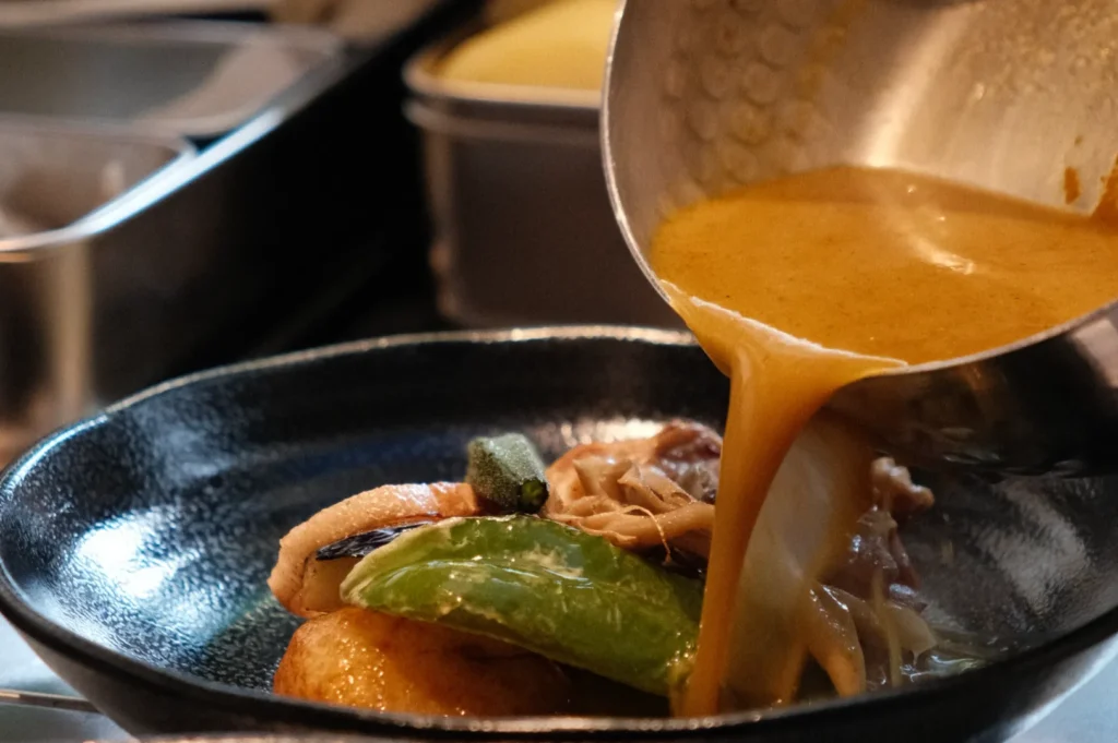 A creamy golden sauce is being poured from a metal ladle over a dish of assorted vegetables in a dark bowl, creating a warm, appetizing scene.