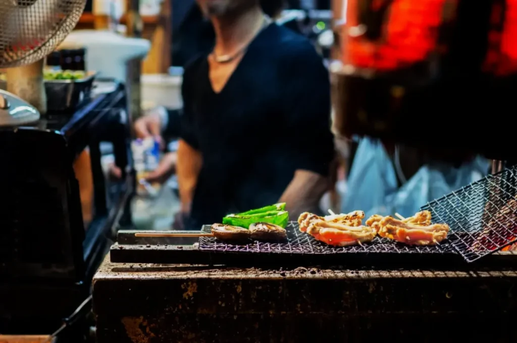 Grilled meat and green vegetables cooking on a street vendor’s charcoal grill with a vendor wearing a black shirt in the blurred background.