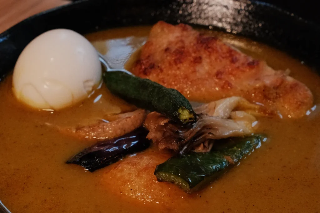 A bowl of curry with a piece of chicken, a soft-boiled egg, and various vegetables such as zucchini and green peppers.