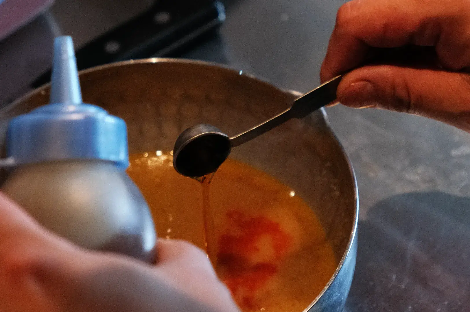 Chef Masa's hands are adding a liquid ingredient from a squeeze bottle into a mixing bowl containing a mixture.