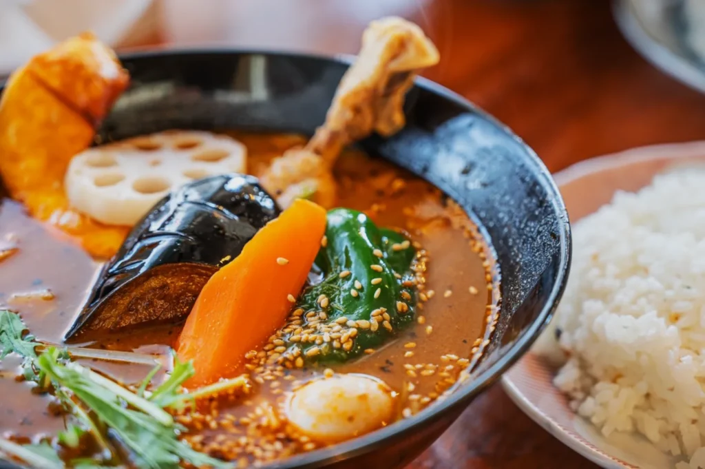 A vibrant bowl of curry soup with chicken, eggplant, carrot, green pepper, and lotus root, topped with sesame seeds. Steaming rice is on the side.
