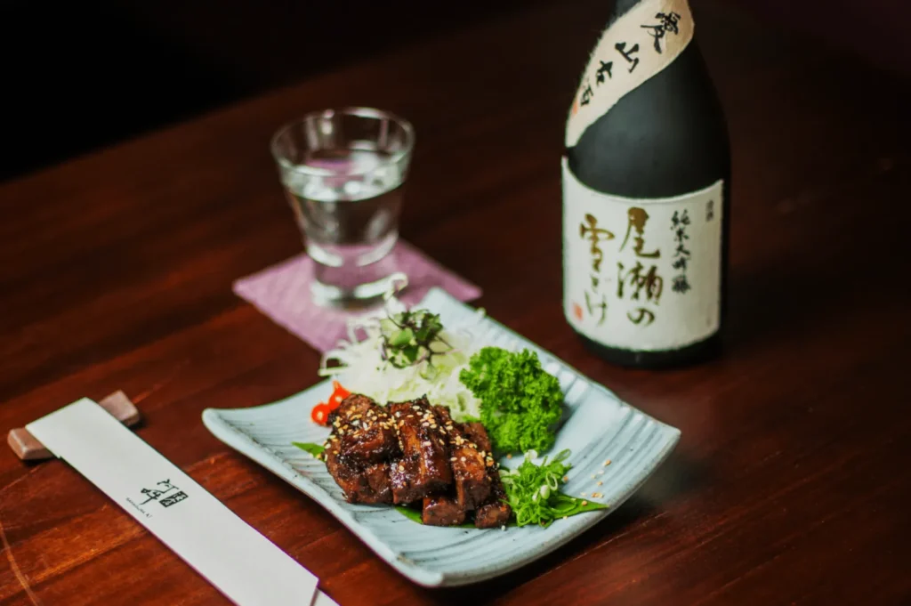 A Japanese meal on a wooden table features grilled meat garnished with sesame seeds, alongside fresh greens. A bottle of sake and a glass are nearby, creating an elegant dining setting.