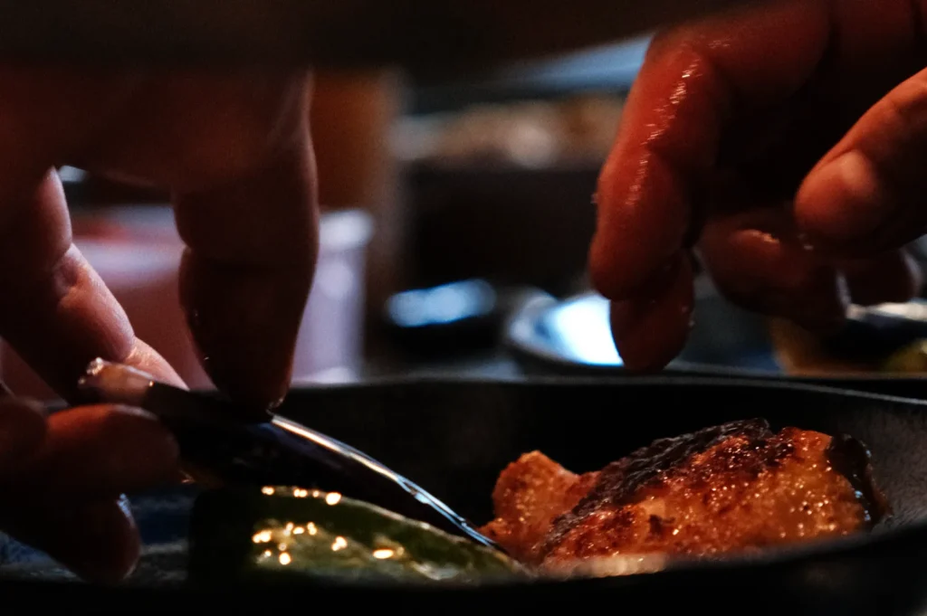 A close-up of hands preparing food in a pan, with one hand holding a spoon and the other reaching for a piece of food.