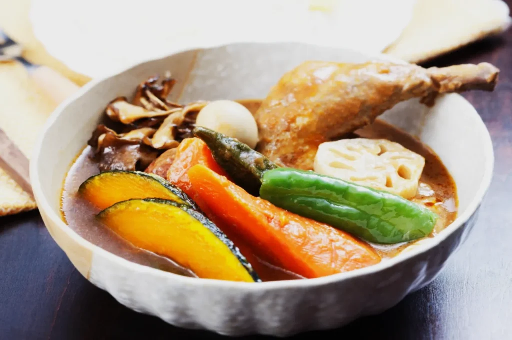 A bowl of Japanese curry soup with a chicken drumstick, lotus root, squash, carrots, green pepper, mushrooms, and a quail egg, conveying warmth.