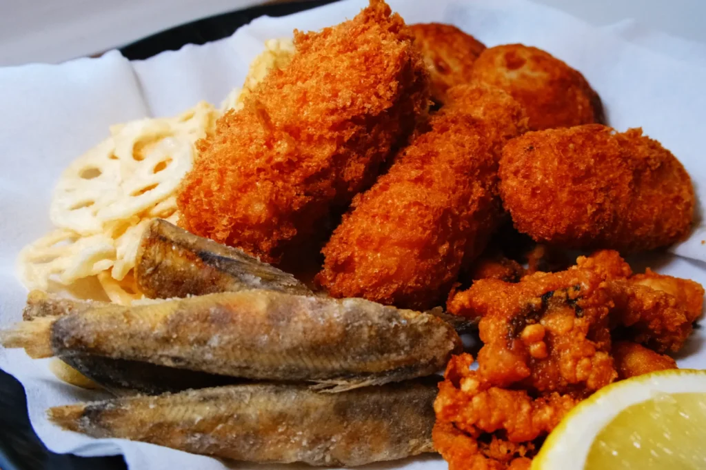 A plate featuring crispy breaded prawns, small fried fish, lotus root chips, and fried chicken, served with a lemon wedge on paper. Enticing and savory.