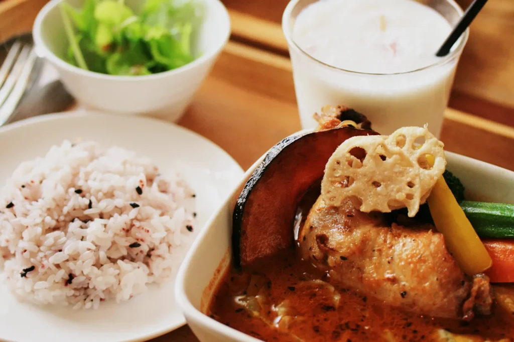 A bowl of creamy soup with chicken, vegetables, and lotus root, alongside a plate of rice, salad, and a frothy drink on a wooden table.