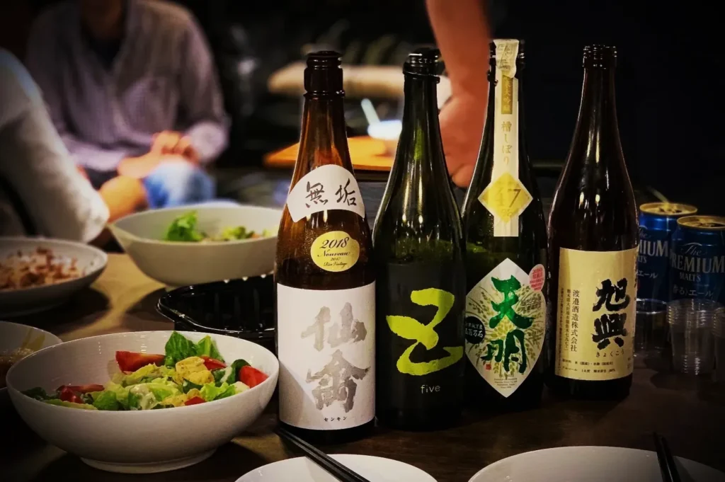 A table with four sake bottles lined up next to bowls of salad. People are seated in the background, suggesting a relaxed gathering.