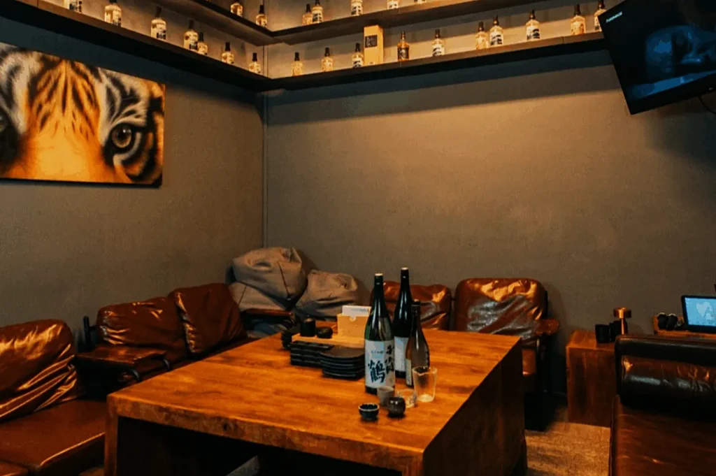 A cozy room with brown leather chairs around a wooden table, topped with bottles and glasses. A tiger painting hangs on the wall; shelves hold small bottles, and a TV is mounted on another wall.