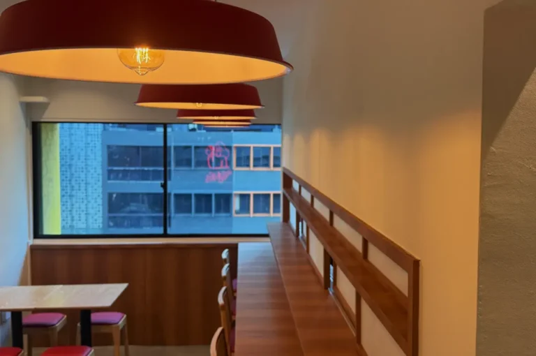 A cozy interior of a restaurant featuring wooden tables and red pendant lights, with a large window showing a city view.