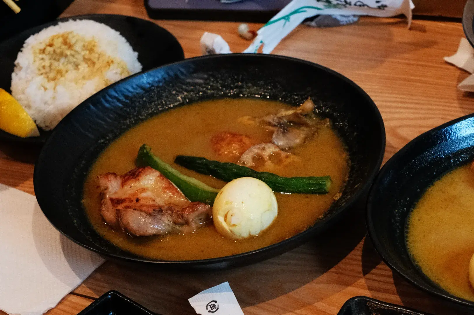 A bowl of soup curry with slices of chicken, green onions, a boiled egg, and a flavorful broth, placed on a wooden table alongside a plate of rice.