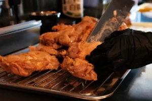 Grilled chicken pieces resting on a rack, with a hand wearing a black glove slicing into one of the pieces.
