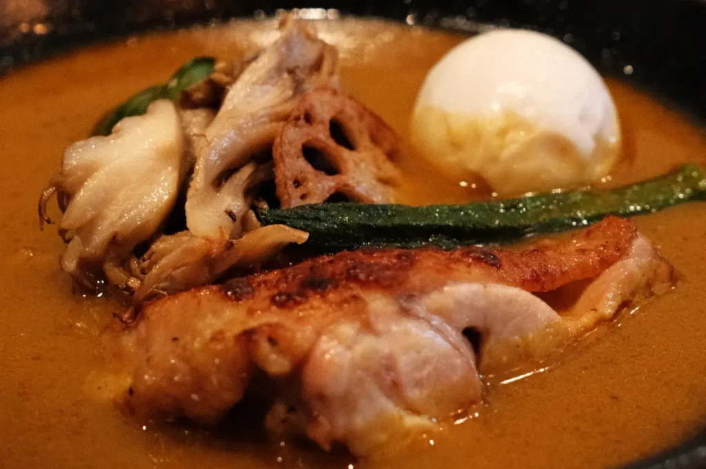 A bowl of rich, flavorful soup with pieces of chicken, mushrooms, lotus root, and a soft-boiled egg, garnished with green vegetables.