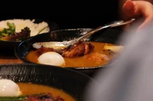 A close-up of a bowl of food featuring meat, an egg, and a sauce, with another plate of rice in the background.