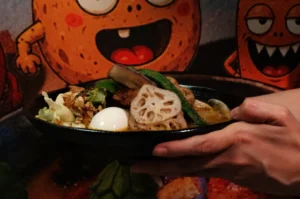 A hand holds a bowl of ramen topped with vegetables, a soft-boiled egg, and a decorative piece of lotus root, with a colorful cartoon background.