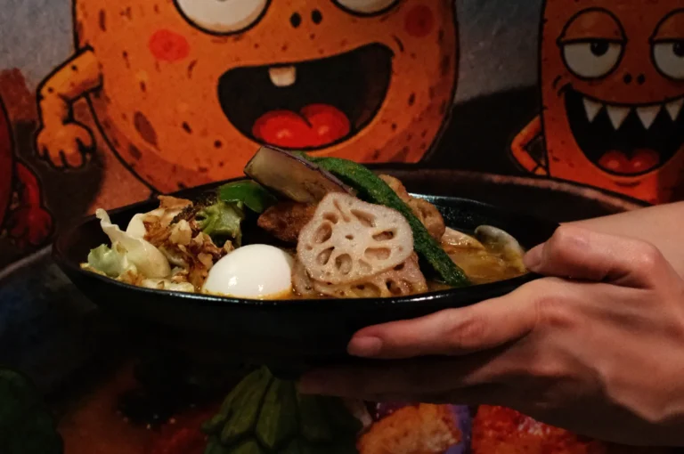 A hand holds a bowl of ramen topped with vegetables, a soft-boiled egg, and a decorative piece of lotus root, with a colorful cartoon background.
