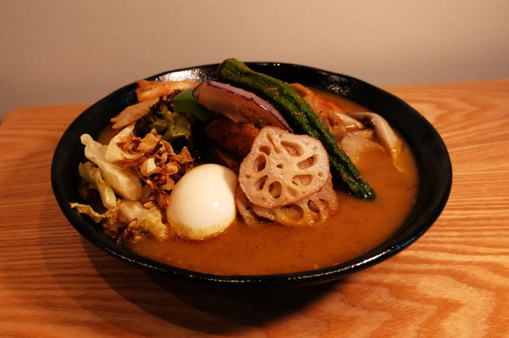 A bowl of soup curry with various toppings, including a soft-boiled egg, lotus root, asparagus, and vegetables in a rich broth.