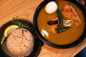 A bowl of soup curry with chicken, an egg, and vegetables, served alongside a plate of rice and a slice of lemon.