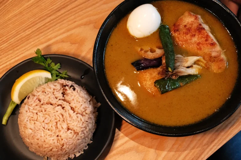 A bowl of soup curry with chicken, an egg, and vegetables, served alongside a plate of rice and a slice of lemon.